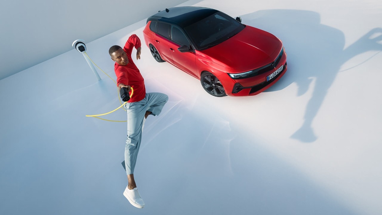 Egy férfi a levegőbe ugrik egy elektromos töltővel, háttérben egy Opel Astra Sports Tourer Electric modellel