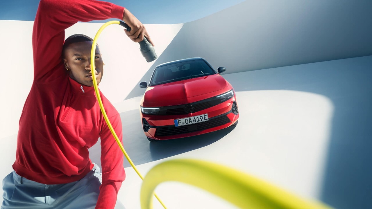 Férfi elektromos töltőkábellel a kezében, egy piros, fekete tetős Opel Astra Sports Tourer előtt áll