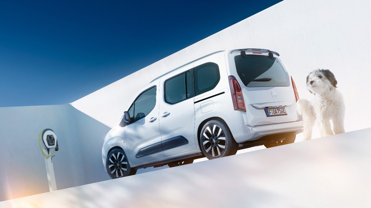 Egy fehér Opel Combo Electric egyterű hátulsó oldalnézetből, és egy kutya egy elektromos töltő mellett